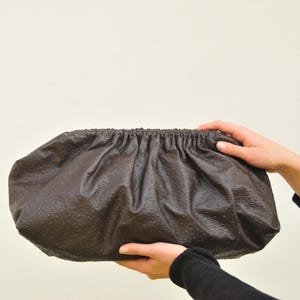 May include: A brown clutch with a gathered top, held by two hands. The clutch has a textured surface with small, evenly spaced bumps. The bag is a dark, warm brown color.