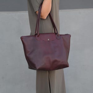 Peut inclure: Un sac cabas en cuir bordeaux foncé doté de deux longues anses, d'un rabat repliable avec un bouton-pression métallique unique et d'un compartiment principal spacieux. Le sac présente une finition lisse et une base structurée avec une large ouverture.