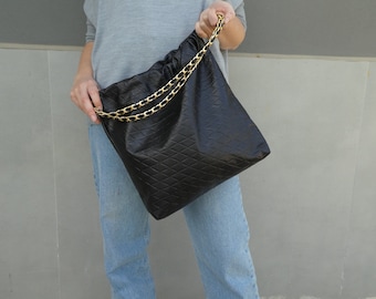 May include: Black quilted handbag with a gold chain strap. The bag has a diamond pattern and a drawstring closure. The chain is a gold color. The bag is held by a person wearing a gray shirt and blue jeans.
