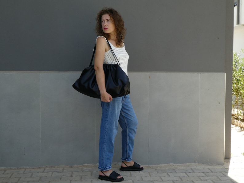 May include: A woman wearing a white tank top and blue jeans is carrying a large black slouchy bag. She is standing in front of a gray wall.