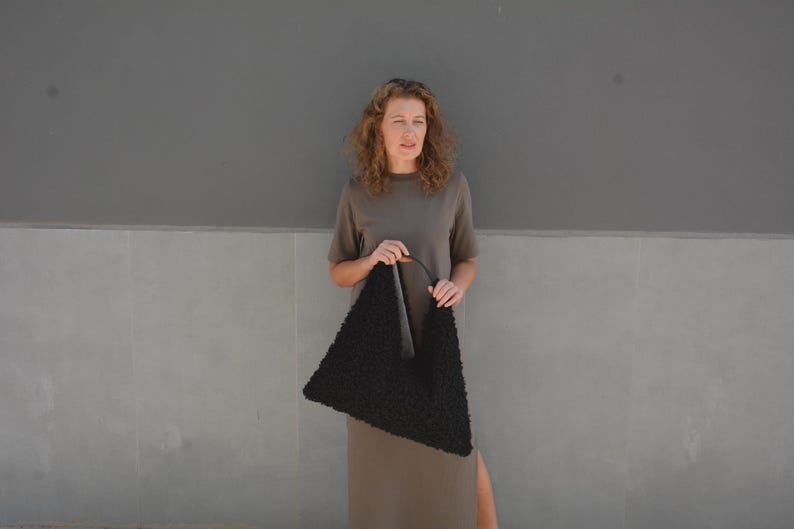 May include: A woman wearing a brown dress holds a black triangular-shaped handbag with a fuzzy texture.