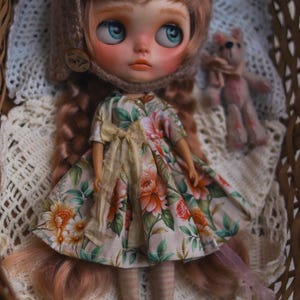 May include: A doll with long braided hair, wearing a beige bonnet and a short-sleeved dress featuring a floral pattern of orange and pink flowers with green leaves. The doll has large blue eyes, rosy cheeks, and wears striped tights with dark shoes.