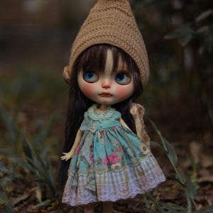 Pode incluir: Uma boneca com cabelos longos e escuros e olhos azuis usa um gorro de crochê bege. A boneca veste um vestido sem mangas azul-petróleo com estampa floral rosa, gola com babados e uma bainha xadrez azul e branca com acabamento em renda.
