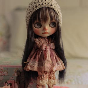 May include: A vintage-style doll with long dark hair and large eyes, wearing a pink dress with lace trim, a crocheted hat, and brown shoes. The doll is standing in front of a floral-patterned box.