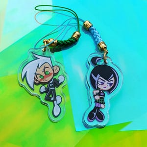 May include: Two cartoon character keychains. One keychain features a character with white hair, green eyes, and a black and white outfit. The other keychain features a character with black hair, purple eyes, and a black and purple outfit. Both keychains have gold-colored hardware and colorful cords.