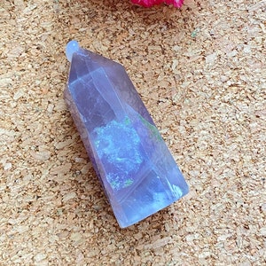 May include: A light purple and blue fluorite crystal point with a pointed top and a flat base. The crystal is sitting on a cork board.