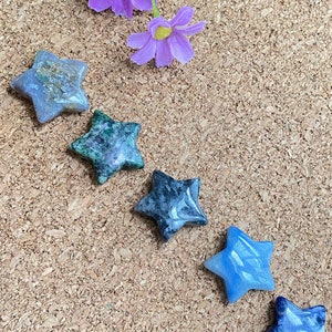 May include: Five star-shaped polished stones in shades of blue and green. The stones are arranged in a row on a corkboard background.