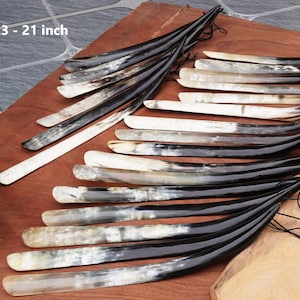 May include: A collection of shoe horns, each with a curved design. The horns are made of a combination of black and cream-colored materials. The text "19.3 - 21 inch" is visible in the image. The shoe horns are arranged on a wooden surface.