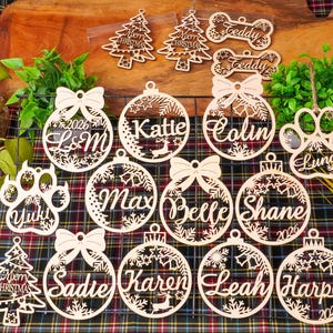 Pode incluir: Uma coleção de ornamentos de madeira cortados a laser com várias formas, incluindo árvores de Natal, ossos de cão, pegadas e esferas circulares. Cada ornamento é personalizado com nomes como Katie, Colin, Max, Belle, Shane, Sadie, Karen, Leah, Harper, Yuki, Luna e Teddy, juntamente com o texto Merry Christmas e o ano 2026.