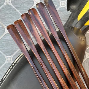 Pode incluir: Um conjunto de seis calçadeiras de madeira castanho-escura com acabamento polido e veios de madeira visíveis. Cada calçadeira está gravada com um nome único em letra cursiva, incluindo Matthew James, Ian Bingham, Thomas Spadaro e Allison L. Farre.