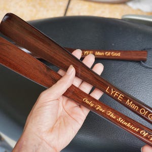 Pode incluir: Duas cal&ccedil;adeiras de madeira castanho-escura com um acabamento liso e polido. A cal&ccedil;adeira superior apresenta o texto gravado LYFE Man Of God, enquanto a inferior exibe o texto gravado Only For The Stinkiest Of Feet em letra cursiva.