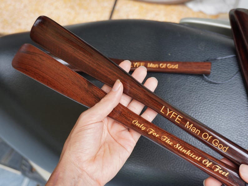 Pode incluir: Duas cal&ccedil;adeiras de madeira castanho-escura com um acabamento liso e polido. A cal&ccedil;adeira superior apresenta o texto gravado LYFE Man Of God, enquanto a inferior exibe o texto gravado Only For The Stinkiest Of Feet em letra cursiva.