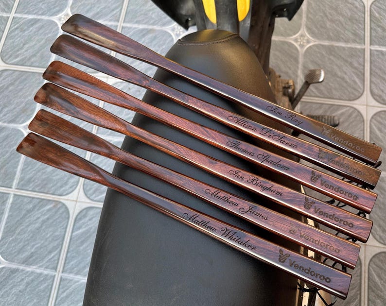 May include: A set of six long, dark brown wooden shoehorns with a polished finish. Each shoehorn features a unique engraved name in cursive script and the brand name Vendoroo with a small rabbit icon printed near the handle end.