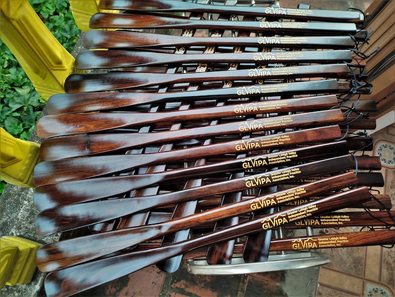 May include: A collection of dark brown wooden shoehorns arranged in a stack. Each shoehorn features a smooth, polished finish and is engraved with the text GLVIPA Greater Lehigh Valley Independent Practice Association, Inc. in gold lettering.