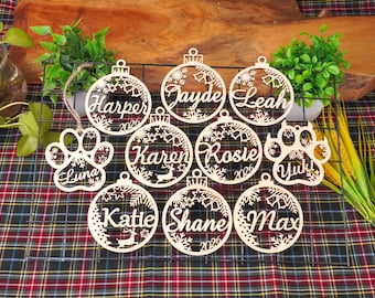 May include: A collection of laser-cut wooden ornaments featuring circular and paw-shaped designs. The ornaments are engraved with names including Harper, Jayde, Leah, Karen, Rosie, Luna, Yuki, Katie, Shane, and Max, along with the year 2026.