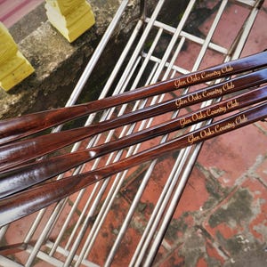 Pode incluir: Quatro cal&ccedil;adeiras longas de madeira castanho-escura dispostas na diagonal. Cada cal&ccedil;adeira apresenta um acabamento liso e polido com o texto Glen Oaks Country Club gravado numa fonte dourada no cabo. Um cord&atilde;o preto fino est&aacute; preso.