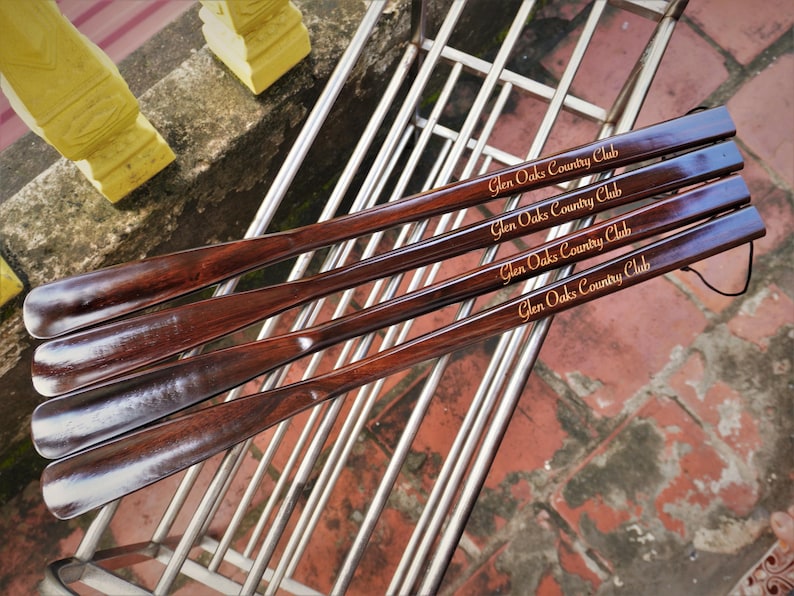 Pode incluir: Quatro cal&ccedil;adeiras longas de madeira castanho-escura dispostas na diagonal. Cada cal&ccedil;adeira apresenta um acabamento liso e polido com o texto Glen Oaks Country Club gravado numa fonte dourada no cabo. Um cord&atilde;o preto fino est&aacute; preso.