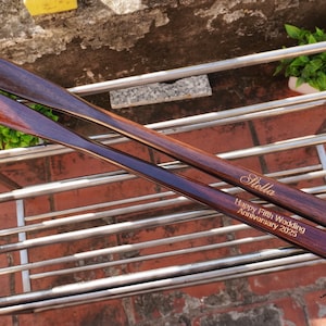 Pode incluir: Duas cal&ccedil;adeiras longas de madeira castanho-escura com um acabamento liso e polido. Uma cal&ccedil;adeira est&aacute; gravada com o nome Stella, e a outra apresenta o texto gravado Happy Fifth Wedding Anniversary 2025 numa fonte clara e leg&iacute;vel.