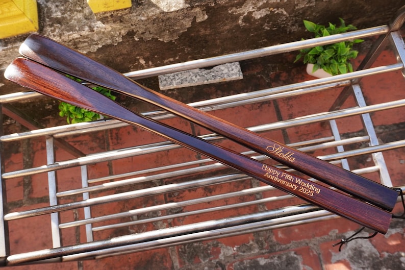 Pode incluir: Duas cal&ccedil;adeiras longas de madeira castanho-escura com um acabamento liso e polido. Uma cal&ccedil;adeira est&aacute; gravada com o nome Stella, e a outra apresenta o texto gravado Happy Fifth Wedding Anniversary 2025 numa fonte clara e leg&iacute;vel.