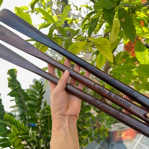 Pode incluir: Tr&ecirc;s cal&ccedil;adeiras longas de madeira castanho-escura com cabos curvos e lisos. Cada cal&ccedil;adeira apresenta um texto gravado a laser: "Owen Max", "Cody & Norma" e "Best man Spen". As superf&iacute;cies de madeira mostram veios naturais e acabamento polido.