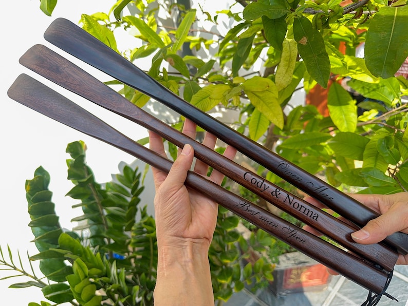 Pode incluir: Tr&ecirc;s cal&ccedil;adeiras longas de madeira castanho-escura com cabos curvos e lisos. Cada cal&ccedil;adeira apresenta um texto gravado a laser: "Owen Max", "Cody & Norma" e "Best man Spen". As superf&iacute;cies de madeira mostram veios naturais e acabamento polido.