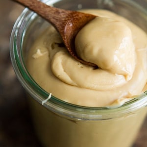 May include: A glass jar filled with a smooth, light brown caramel sauce. A wooden spoon is scooping out the sauce, showing its thick, creamy texture.