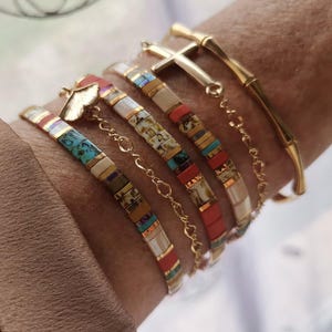 Könnte beinhalten: Eine Sammlung farbenfroher Armbänder mit Perlen und ein goldener Bambus-Armreif. Die Armbänder weisen eine Vielzahl von Farben auf, darunter Türkis, Rot und Creme, mit goldenen Akzenten. Ein Armband hat einen goldenen Kreuzanhänger.