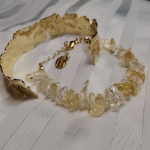 Peut inclure: Un bracelet doré avec un bord texturé et irrégulier. Le bracelet présente une section de pierres de citrine claires et jaune. Le bracelet est présenté sur une surface blanche et rayée.