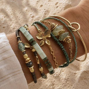 Peut inclure: Un ensemble de cinq bracelets superposés comprenant un jonc en métal doré avec un nœud, des brins de perles vertes, une breloque papillon en métal doré et un bracelet composé de tuiles rectangulaires vert pâle et vert foncé avec des détails dorés.
