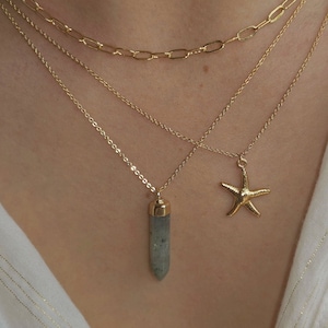 Könnte beinhalten: Ein mehrschichtiges Halsketten-Set mit drei goldenen Ketten. Die obere Kette hat große rechteckige Glieder, die mittlere Kette ist filigran und die unteren Ketten haben einen Kristallanhänger und einen Seesternanhänger. Die Halsketten werden auf weißem Hintergrund präsentiert.
