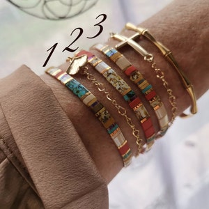 May include: A collection of gold bracelets and beaded bracelets. The bracelets include a gold cross charm, a bamboo-style bracelet, and several bracelets with colorful rectangular beads. The image has the numbers 1, 2, and 3 in the upper left corner.