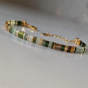 Può includere: Un braccialetto delicato con una catena dorata e una sezione di perline rettangolari verdi, marroni e trasparenti. Il design del braccialetto è sottile ed elegante, adatto per l'uso quotidiano o per occasioni speciali.