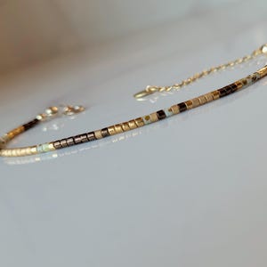 Op de afbeelding: Delicate armband met kleine, vierkante kralen in goud, bruin en crème. De armband heeft een gouden ketting en sluiting. De kralen zijn in een lineair patroon gerangschikt, wat een minimalistisch en elegant ontwerp creëert.
