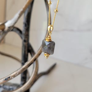 Puede incluir: Un collar dorado con un colgante cuadrado de piedra gris. El colgante está engastado en filigrana dorada y cuelga de una fina cadena con pequeñas cuentas doradas. El collar se muestra sobre un fondo borroso.