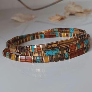 Könnte beinhalten: Ein mehrreihiges Armband in Gold-, Türkis-, Orange- und Brauntönen. Das Armband besteht aus kleinen, rechteckigen Perlen und goldfarbenem Metall. Das Armband ist gewickelt und zeigt sein aufwendiges Design. Der Umfang beträgt ca. 18 cm.
