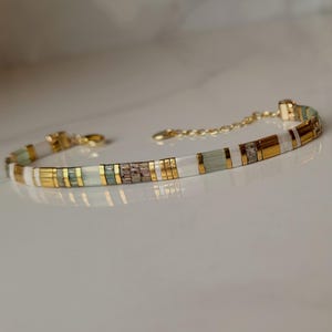 Puede incluir: Una pulsera delicada con un patr&oacute;n repetitivo de cuentas cil&iacute;ndricas en oro met&aacute;lico, blanco opaco y verde salvia transl&uacute;cido. El dise&ntilde;o incluye una cadena extensora de metal dorado con un cierre de mosquet&oacute;n para un ajuste regulable.
