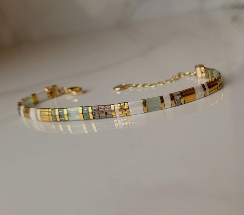 Puede incluir: Una pulsera delicada con un patr&oacute;n repetitivo de cuentas cil&iacute;ndricas en oro met&aacute;lico, blanco opaco y verde salvia transl&uacute;cido. El dise&ntilde;o incluye una cadena extensora de metal dorado con un cierre de mosquet&oacute;n para un ajuste regulable.