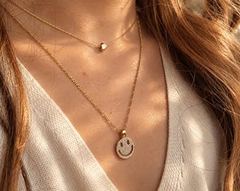 May include: A layered gold necklace set featuring two chains. The shorter chain has a small, smooth gold heart charm. The longer chain features a circular pendant with a smiley face design, fully encrusted with small, clear, sparkling stones.