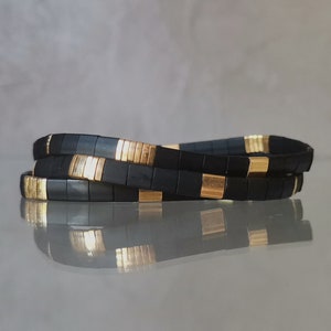 May include: Three black and gold tile bracelets are stacked on a reflective surface. The bracelets are made of small, square black tiles with gold-coloured accents. The bracelets are coiled, showing their flexibility.