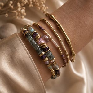 Peut inclure: Un empilement de quatre bracelets comprenant un jonc doré style bambou, un fin rang de perles de rocaille blanches et dorées, un rang de perles violettes et dorées, et un bracelet multi-rangs avec des perles facettées grises et violettes.