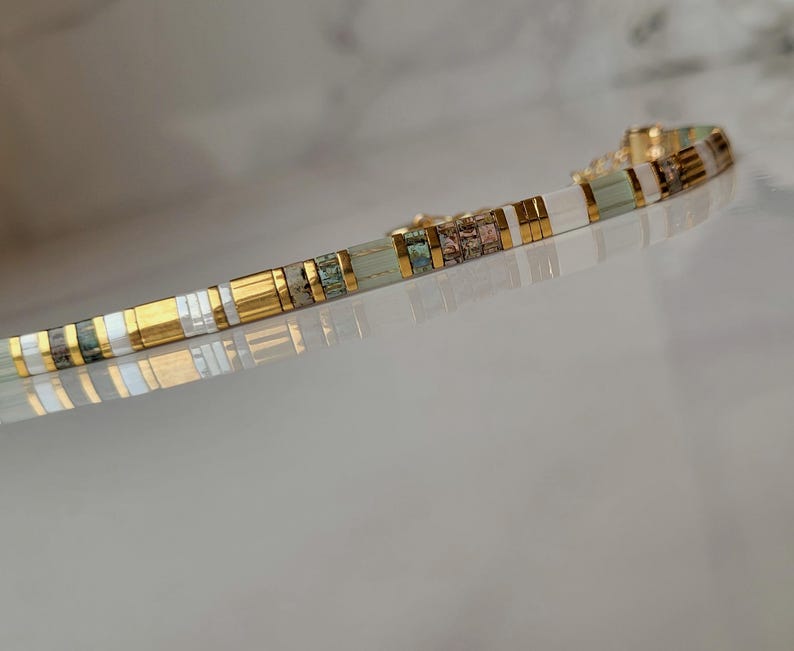 Puede incluir: Una pulsera delicada compuesta por cuentas rectangulares alternas en tonos verde salvia, blanco y oro met&aacute;lico. El dise&ntilde;o presenta un patr&oacute;n repetitivo de segmentos lisos de colores s&oacute;lidos intercalados con peque&ntilde;as cuentas texturizadas.