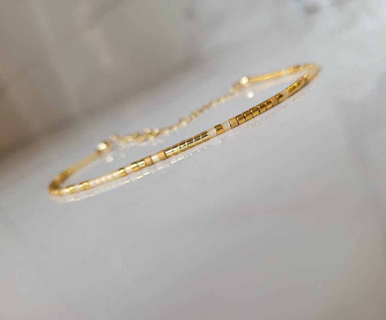 Op de afbeelding: Delicate goudkleurige armband met een reeks kleine, rechthoekige kralen in goud en gebroken wit. De armband heeft een fijne ketting en sluiting. Het ontwerp is eenvoudig en elegant, geschikt voor dagelijks gebruik of speciale gelegenheden.