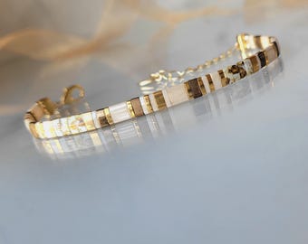 May include: A delicate bracelet composed of rectangular beads in a palette of cream, metallic gold, dark brown, and white with speckled patterns. The piece features a gold-toned metal lobster clasp and an adjustable chain extension.