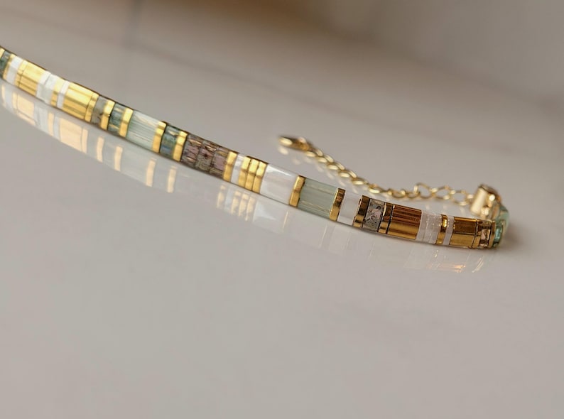 Puede incluir: Una pulsera delicada compuesta por cuentas rectangulares alternas en oro met&aacute;lico, blanco opaco, verde p&aacute;lido transl&uacute;cido y un patr&oacute;n gris-marr&oacute;n moteado. La pieza presenta una cadena extensora de metal dorado con un peque&ntilde;o cierre al final.