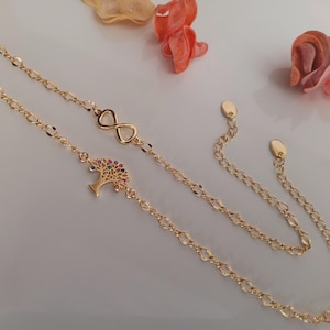 May include: A gold-toned chain bracelet featuring an infinity symbol and a tree of life charm adorned with small, multi-coloured gemstones. The bracelet includes a delicate link chain and an adjustable extender with two small, flat, oval-shaped end tags.