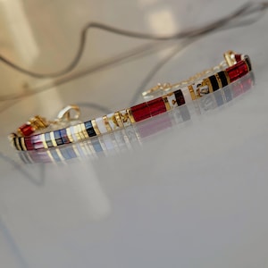 Puede incluir: Una pulsera dorada con segmentos rectangulares rojos, blancos, negros y dorados. La pulsera tiene una cadena y un cierre delicados. El diseño incorpora un patrón repetitivo de colores y formas, creando un accesorio elegante.