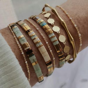 Peut inclure: Plusieurs bracelets dorés et perlés sont présentés. Les bracelets comprennent des chaînes en or, des breloques en forme de disques et des éléments en forme de bambou. Les bracelets de perles présentent des perles dorées, vertes et crème.