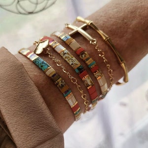May include: A collection of colorful bracelets and a gold bangle are displayed on a wrist. The bracelets feature a variety of colors including turquoise, red, and cream. One bracelet has a gold butterfly charm, and another has a gold cross charm.