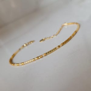 Op de afbeelding: Delicate gouden armband met kleine, rechthoekige kralen in goud, cr&egrave;me en bruin. De armband heeft een verstelbare ketting en sluiting. De sieraden worden gepresenteerd op een wit oppervlak.