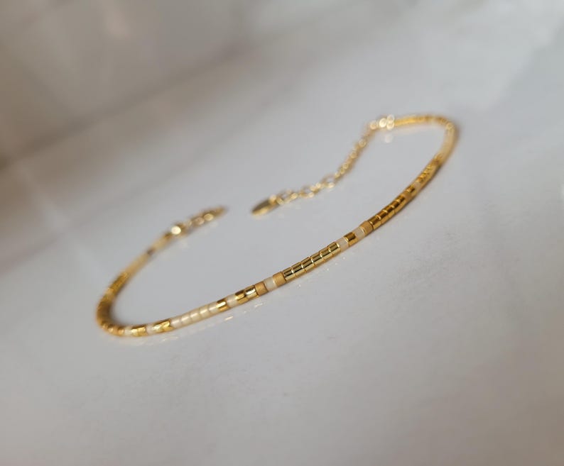 Op de afbeelding: Delicate gouden armband met kleine, rechthoekige kralen in goud, cr&egrave;me en bruin. De armband heeft een verstelbare ketting en sluiting. De sieraden worden gepresenteerd op een wit oppervlak.